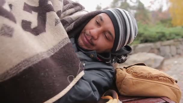 Несчастный человек лежит на скамейке, смотрит в камеру с грустными глазами, страдает от холода — стоковое видео