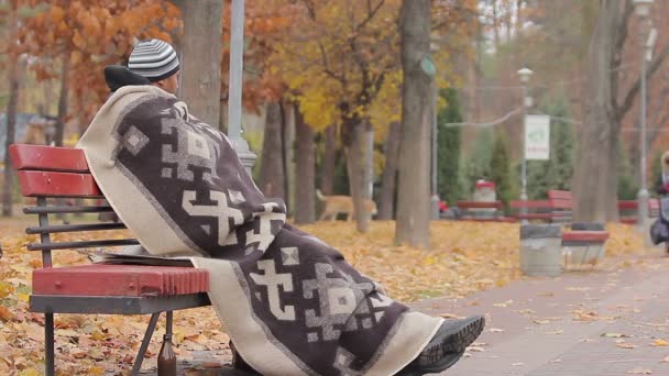 Бездомный сидит на скамейке с грустным лицом, смотрит на людей в осеннем парке — стоковое видео