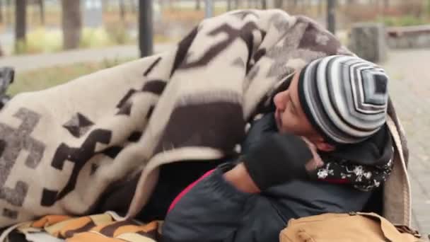 冷冻的男子躺在公园里，坐在板凳上把自己裹在毯子，贫穷 — 图库视频影像