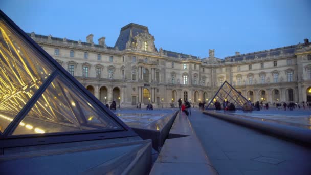 Mucha gente caminando en el patio del Palacio del Louvre, viendo construcciones piramidales de vidrio — Vídeo de stock