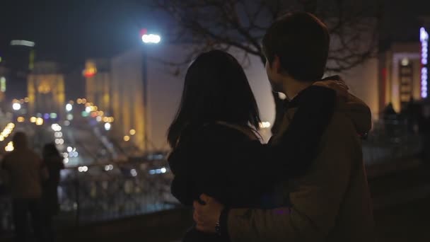 Amar joven hombre y mujer abrazándose suavemente, mirando románticas luces de la ciudad de noche — Vídeo de stock