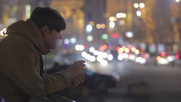 Young lonely man drinking hot tea on street, watching night city, sadness — Stock Video