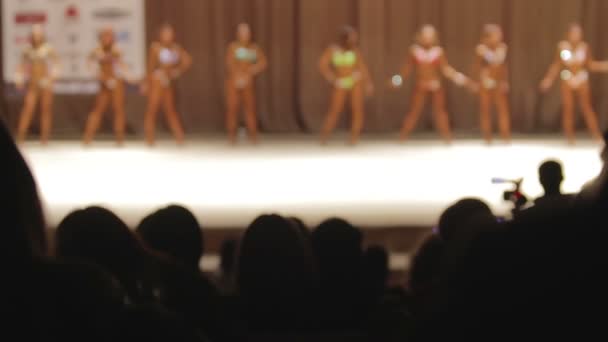 Pessoas assistindo competição figura fitness, mulheres de biquíni se apresentando no palco — Vídeo de Stock