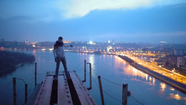 Счастливый человек стоит на вершине моста и указывает на прекрасный вид на ночной город — стоковое видео