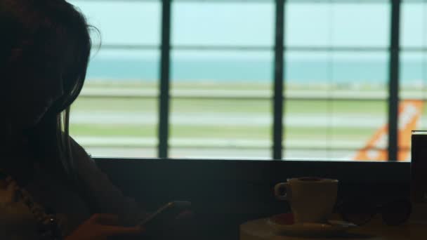 Mooi meisje chatten met vriendje op de telefoon, drinken koffie in het café van de luchthaven — Stockvideo