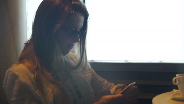 Junge Frau mit Sonnenbrille, die im Café auf dem Smartphone SMS schreibt und im Internet surft — Stockvideo