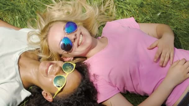 Dos hermosas mujeres tumbadas en el césped verde, hablando, disfrutando de las vacaciones al aire libre — Vídeos de Stock