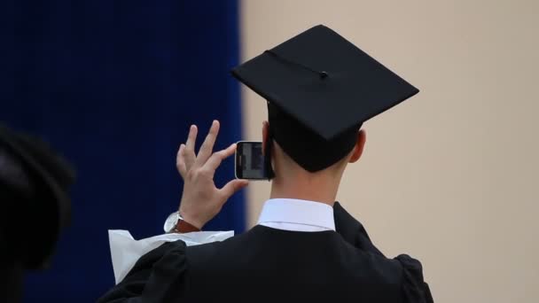 Jovem em vestido acadêmico e chapéu fotografando cerimônia de formatura no telefone — Vídeo de Stock