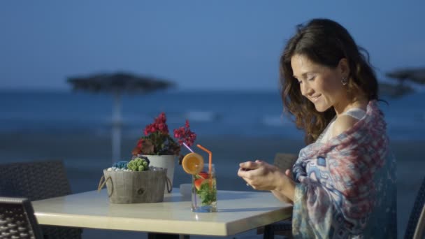 Mulher bonita mensagens de texto no telefone móvel com sorriso no rosto, descanso no café à beira-mar — Vídeo de Stock