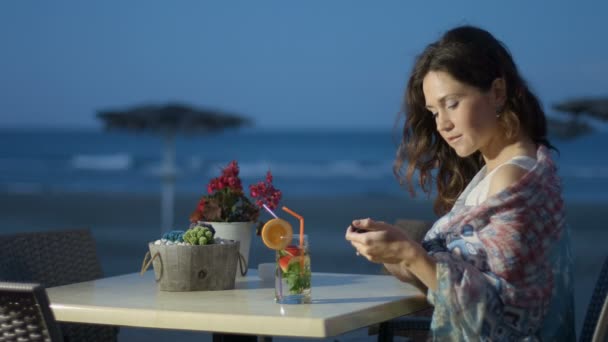 बीच रेस्टॉरंटमध्ये विश्रांतीचा आनंद घेणारी रोमँटिक स्त्री, फोनवरून संदेश पाठविते — स्टॉक व्हिडिओ