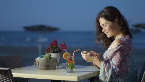 Hermosa joven utilizando el teléfono móvil en el restaurante al aire libre, mirando a la cámara — Vídeos de Stock
