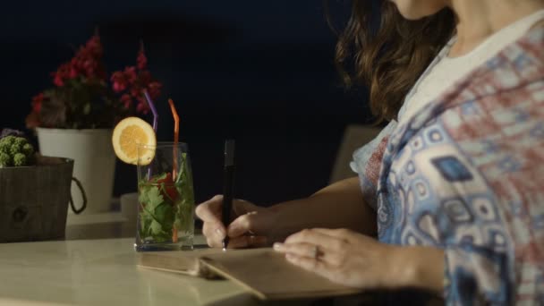 Manos de mujer inspirada escribiendo poesía en bloc de notas en un restaurante al aire libre por la noche — Vídeos de Stock