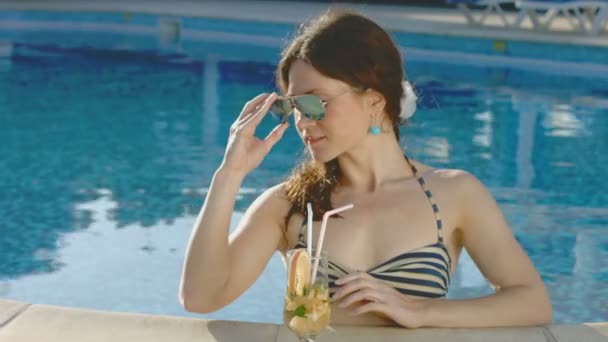 Красивая модель пьет коктейль в бассейне, смотрит в камеру — стоковое видео