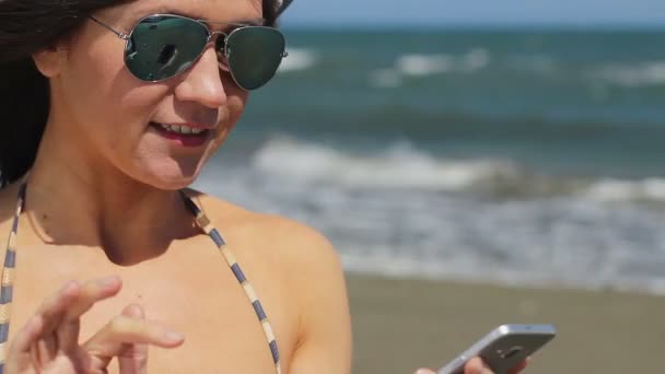 Mujer joven en gafas de sol respondiendo a una llamada telefónica en la playa, sonriendo, hablando — Vídeo de stock