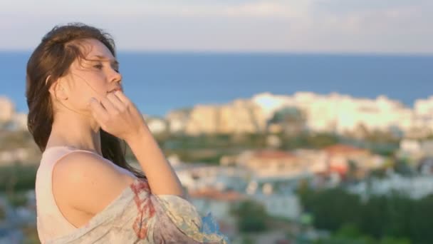 Красивая женщина стоит на холме и смотрит на красивый морской город. Волосы машут ветром — стоковое видео