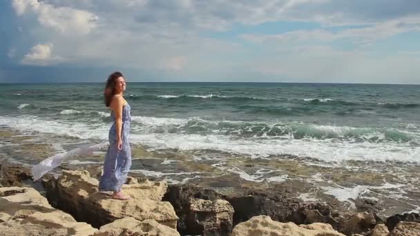 Feliz joven mujer relajándose en la playa rocosa en tiempo ventoso, mirando a la cámara — Vídeo de stock