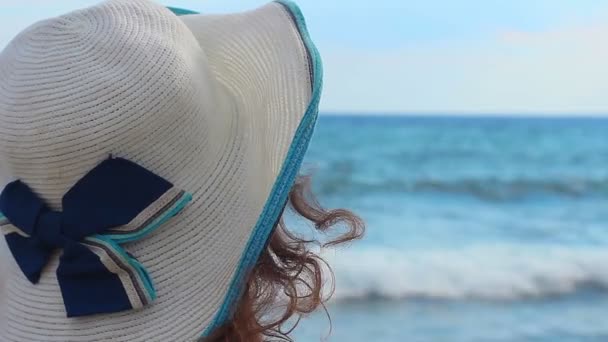 Visão traseira de uma jovem triste de chapéu sentada sozinha na praia, olhando para as ondas do mar — Vídeo de Stock