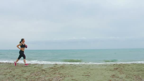 Schönes Mädchen joggt am Meer entlang. Salzige Wellen plätschern am leeren Sandstrand — Stockvideo