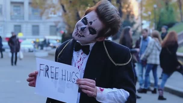 Молодой человек в костюме зомби держит табличку с объятиями в руках, улыбаясь в камеру — стоковое видео