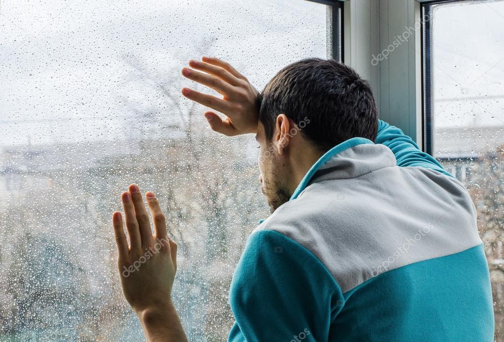 Depressed young man upset with bad news looking through rainy window glass