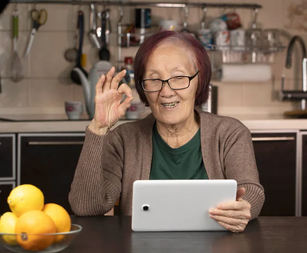 Elderly asian woman hand gesture okay and holding digital tablet computer video conference with family in the kitchen during quarantine. Elderly people use technology concept.