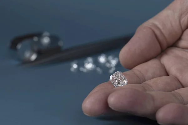 Cut diamond in hand close up with jewelry tools and scattering of different diamonds in background, front view on blue background. — Stock Photo, Image