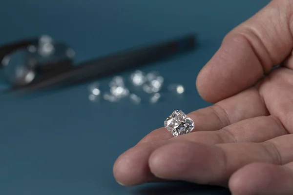 Vágott gyémánt a kézben, közel az ékszerek szerszámok és szórása a különböző gyémántok a háttérben, elölnézet a kék háttér. — Stock Fotó