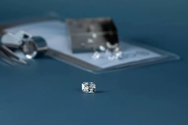 Diamante tallado en mano de cerca con herramientas de joyería y la dispersión de diferentes diamantes en el fondo, vista frontal sobre fondo azul. — Foto de Stock