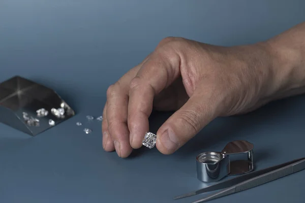 Cut diamond in hand close up with jewelry tools and scattering of different diamonds in background, front view on blue background.