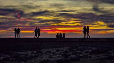 Faro de Maspalomas deniz feneri, ziyaretçiler gezinti güvertesi ve Atlantik manzarasının keyfini çıkarırken parlayan bir alacakaranlık gökyüzüne karşı dimdik ayakta duruyor. İnsanların siluetleri ve renkli ufuk sakin ama canlı bir kıyı atmosferi yaratır..