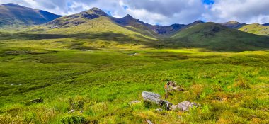 Iskoç Dağları 'ndaki sarp dağlar arasında yemyeşil bir vadi esiyor, dramatik bulutlar ve yumuşak ışık uzak manzaranın doğal dokularını ve sakin atmosferini güçlendiriyor..