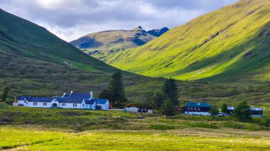 İskoçya 'nın yüksek kesimlerinde sarp yeşil tepelerle çevrili ve beyaz kulübelerle benekli pitoresk bir vadi. Güneş ışığı ve bulutlar, bu uzak ortamın doğal hatlarını ve barışçıl cazibesini vurgulayarak, manzara boyunca dramatik gölgeler oluşturuyor.