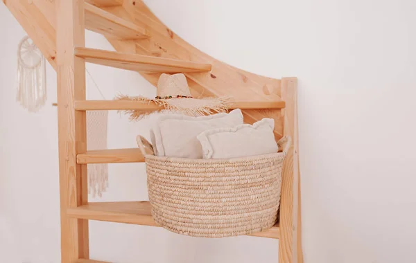 wicker storage basket with white pillow inside on wooden spiral staircase against white clean wall. stylish and convenient system storage at home