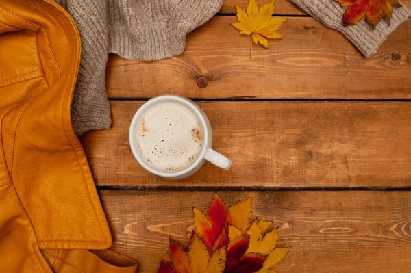 cozy autumn flatlay. Cup of coffee with milk foam, beige knitted sweater, yellow leather jacket and red maple leaves on rough wooden table, selective focus