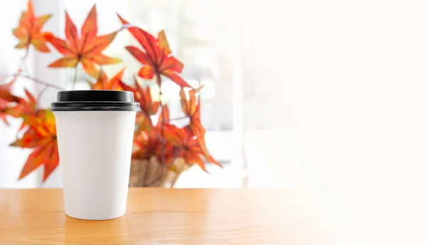 white paper Cup with black lid for hot drinks on wooden table in cafe on background of red and orange autumn leaves by the window, banner with space for text, selective focus