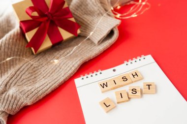 Kırmızı arkaplanda beyaz sarmal defter ve kare harflerle dilek listesi, arka planda kırmızı saten fiyonklu Kraft kağıdından yapılmış hediye kutusu, çelenkler ve örülmüş kumaş.