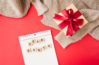 white spiral notebook and square inscription dear Santa on red background, gift box made of Kraft paper with red satin bow, garlands and knitted fabric on background, selective focus