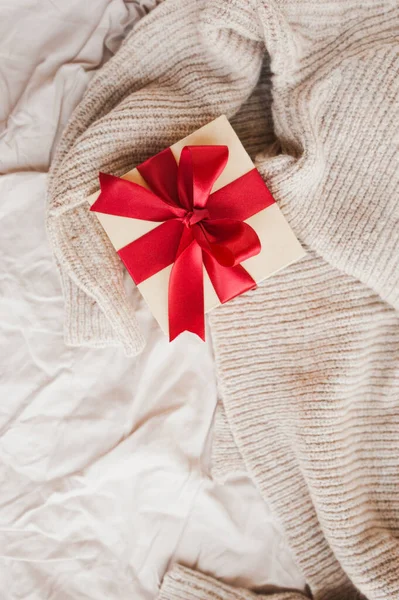 stylish square beige box with red festive satin ribbon on beige cozy crumpled blanket selective focus