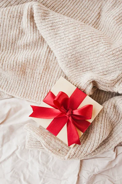 stylish square beige box with red festive satin ribbon on beige cozy crumpled blanket selective focus