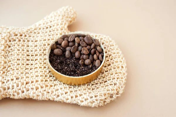 jar with coffee scrub and grains on rough massage brush glove made of jute on clean beige background, cosmetic layout home body care, selective focus