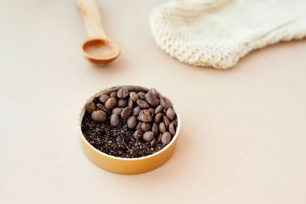 Coffee scrub large granules and coffee beans in gold jar, cosmetic oil in wooden spoon and rough massage glove against pastel beige background, homemade skincare, selective focus