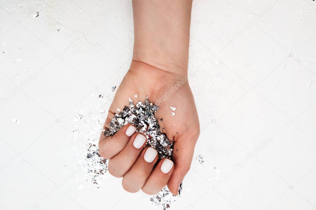 light-skinned hand with perfect manicure with milky color gel polish holds large handful of silver foil sequins on white background, selective focus