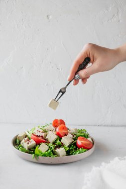 İki çatallı el, tofu peyniri salatasına uzanıyor, otlar ve domatesler beyaz masada. dikey yiyecek içeriği keto diyeti, seçici odaklanma