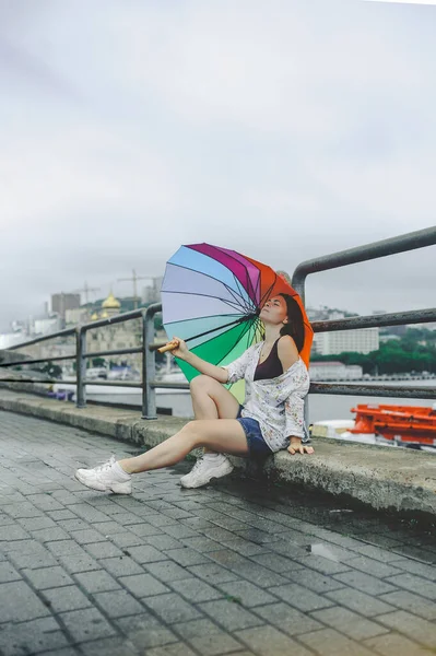 young beautiful woman in floral print shirt, denim shorts and white large sneakers sits on wet paving stones in rainy weather under rainbow umbrella in an urban atmosphere, vertical content