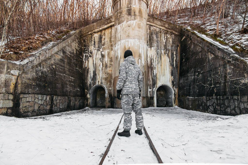 man in warm military uniform,black hat and tactical gloves stands at tall old fortress with narrow gauge railway in winter, selective focus