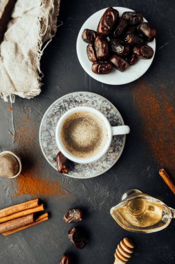 Doğu yemeği. Taze demlenmiş kahve, kurutulmuş hurma, tarçın çubukları, bal dökülmüş kahve ve baharatlar, koyu arkaplanda çuval bezi. Ulusal gıda ve içeceğin renkli görüntüsü. Üst görünüm, seçici odak