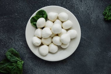 Beyaz porselen tabakta küçük mozzarella peyniri topları ve siyah masada taze yeşil yapraklar. Peynir türleri, evde peynir pişirme tarifleri. şık yemek içeriği, seçici odaklanma