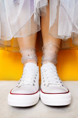 Beyaz, temiz, yeni spor ayakkabılı kadın bacakları şeffaf, parlak gümüş renkli çoraplar ve sarı arka planda gür tül elbiseler. Spor ayakkabıları ve resmi kıyafetlerin birleşimi. seçici odak