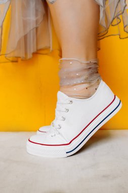 Beyaz, temiz, yeni spor ayakkabılı kadın bacakları şeffaf, parlak gümüş renkli çoraplar ve sarı arka planda gür tül elbiseler. Spor ayakkabıları ve resmi kıyafetlerin birleşimi. seçici odak