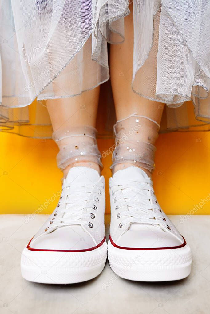 women legs in white clean new sneakers, transparent thin socks with silver shiny stars and lush tulle dress on yellow background. combination of sports shoes and formal wear. selective focus